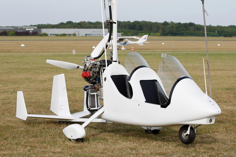A white tandem two-seat gyroplane parked on grass at an airfield, seen from the side, with its open cockpit, pusher propeller and twin tail fins visible and a light aircraft in the background.