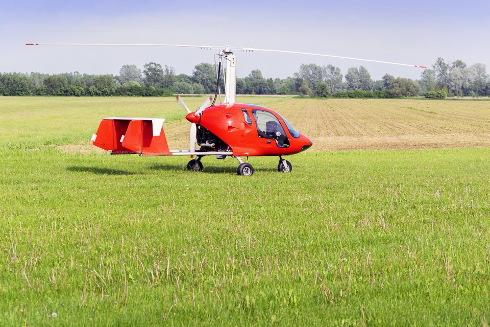A red enclosed-cabin gyroplane parked on a grass field with a treeline behind it, rotor blades extended and twin red tail fins.