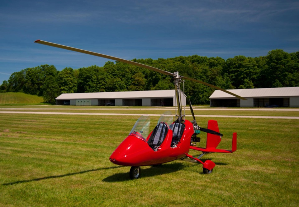 A red open-cockpit tandem two-seat gyroplane parked on mown grass at an airfield, with a row of white hangars and a treeline behind it.