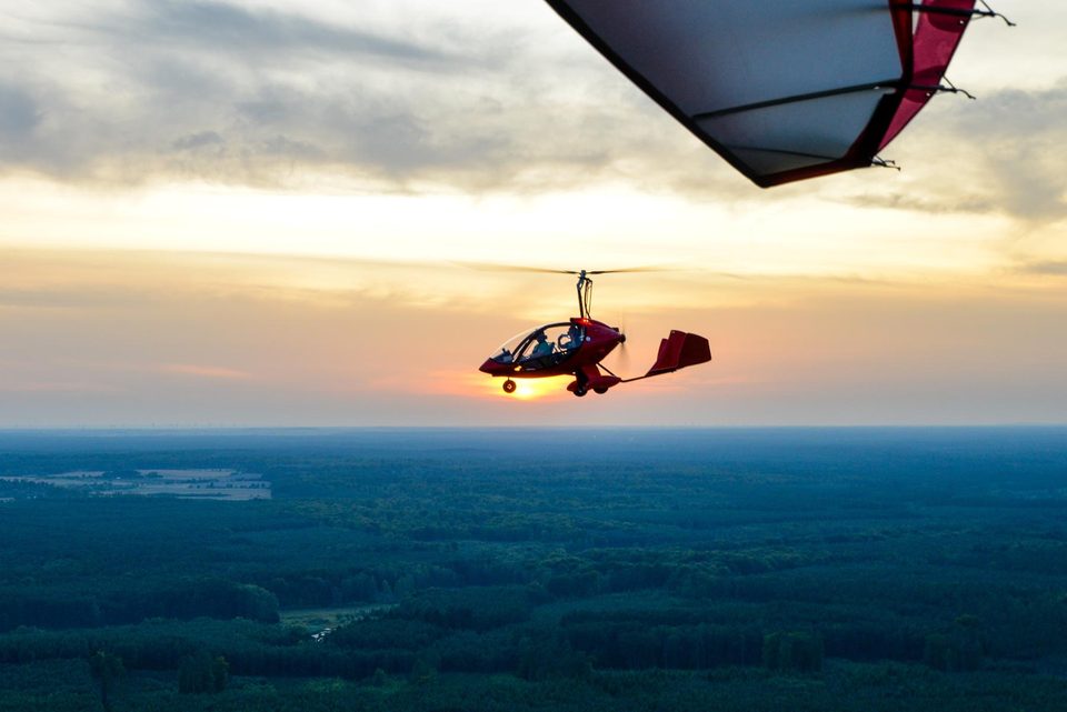 A red two-seat gyroplane in flight above forest at sunset, photographed from another aircraft whose wing edge is visible at the top of the frame.