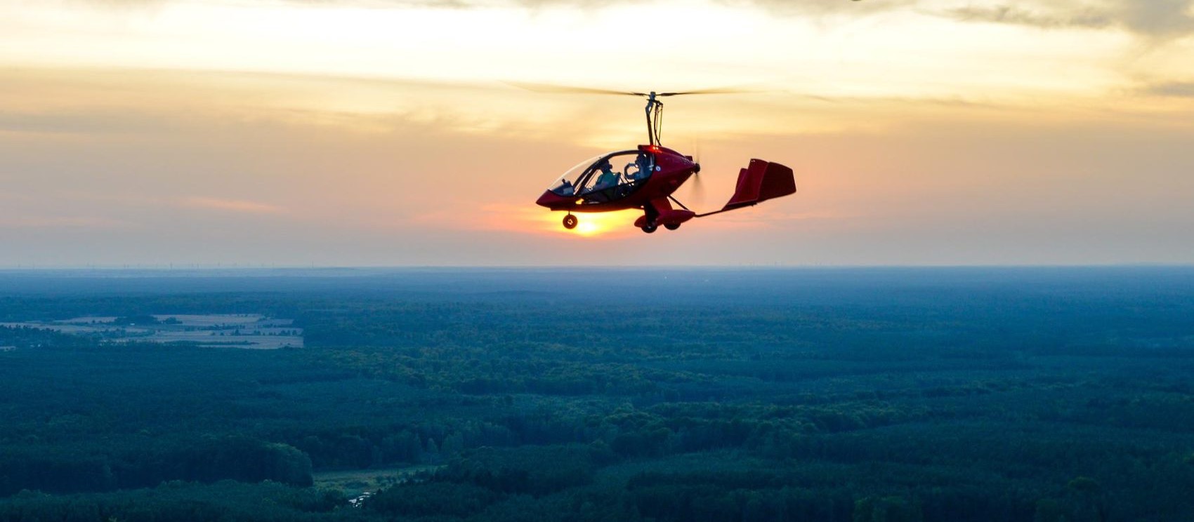 Red gyroplane in flight over forest at sunset