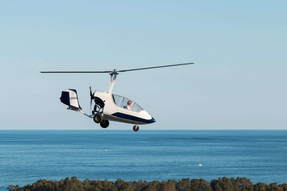 A white gyroplane in flight over open water.