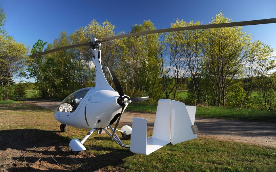 A white gyrocopter parked at the edge of a woodland trail, rotor and tail in full view.